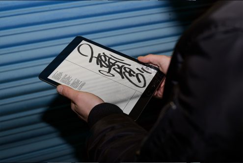 Person holding a tablet showcasing calligraphy font mockup with graffiti style on a digital screen, set against a blurred blue background.