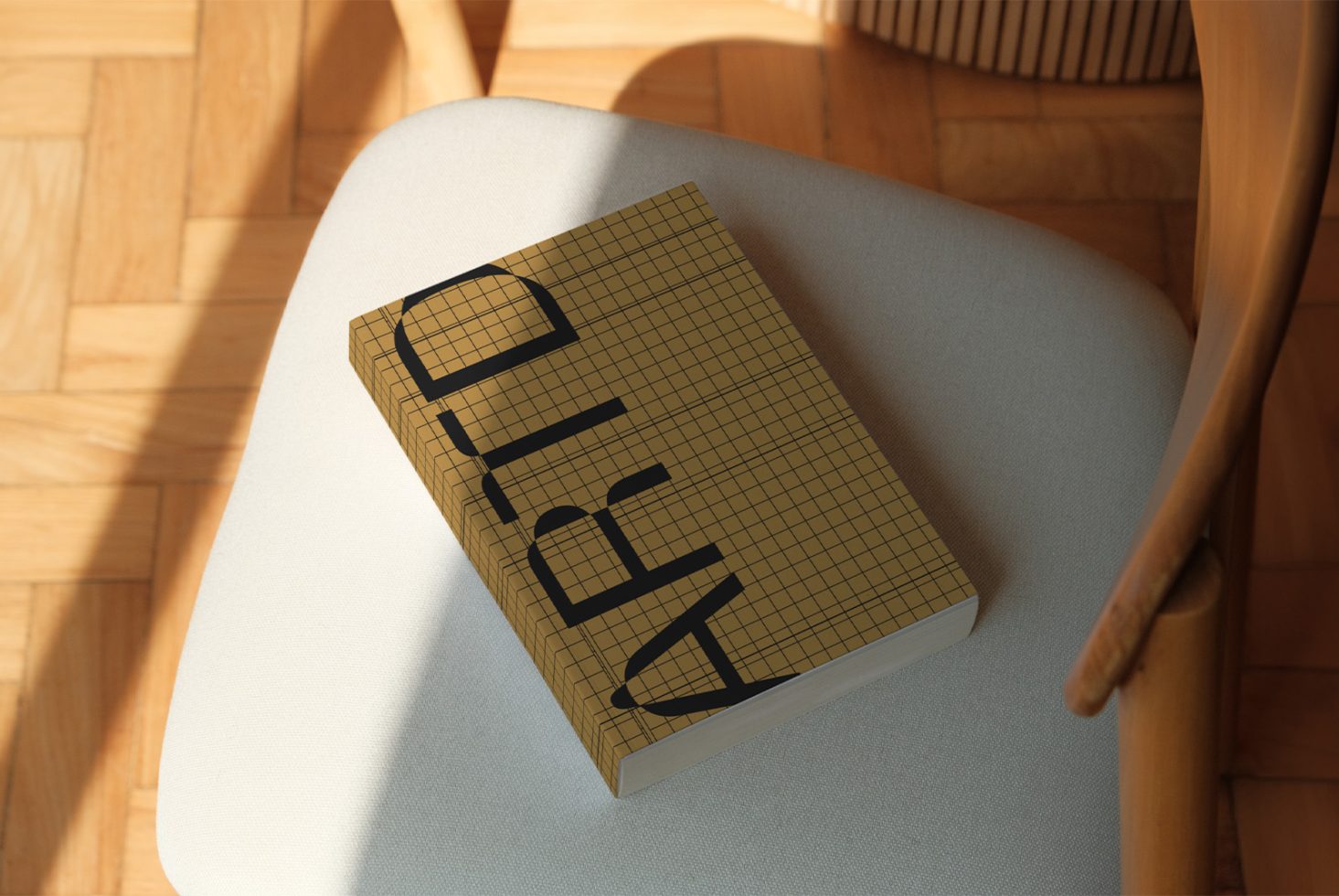 Contemporary notebook mockup on chair, grid paper texture cover with bold typography, natural light, wooden floor background.