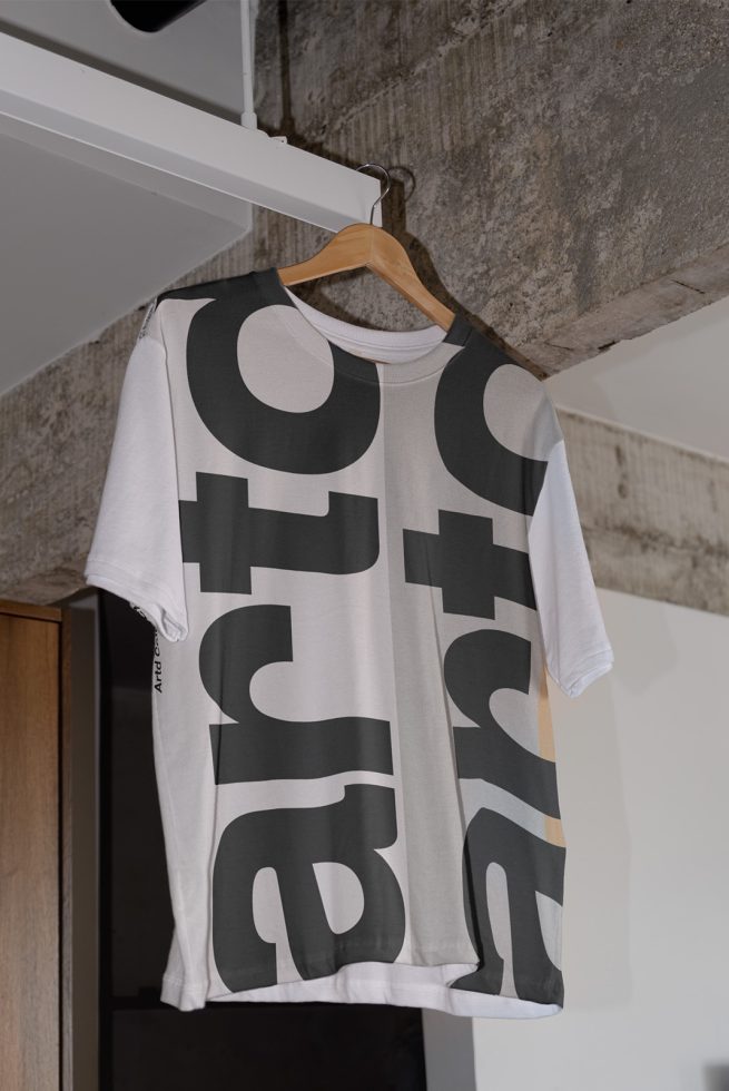 White t-shirt mockup hanging on wooden hanger against a concrete wall, featuring bold black typography design, ideal for fashion and apparel graphics.