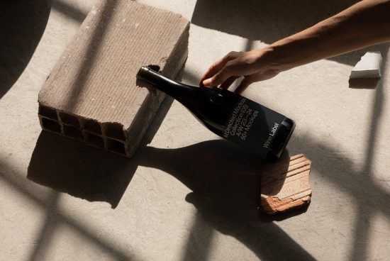 Hand positioning black wine bottle on concrete with shadows for product mockup design, elegant presentation, wine label showcase.