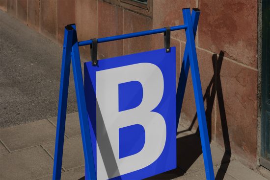 Blue sidewalk signboard mockup with a bold white letter B, standing on a pavement against a warm-hued wall, clear shadows, and daylight.