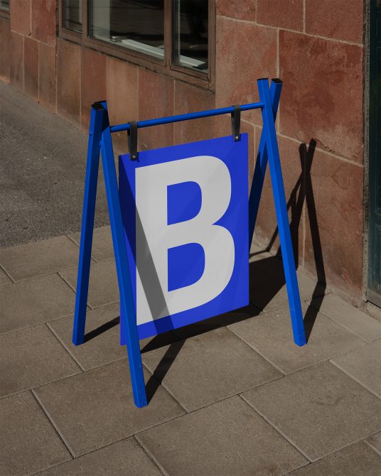 Blue A-frame sidewalk sign mockup with a bold white letter B, standing on a pavement against a brick wall, suitable for font display in urban settings.