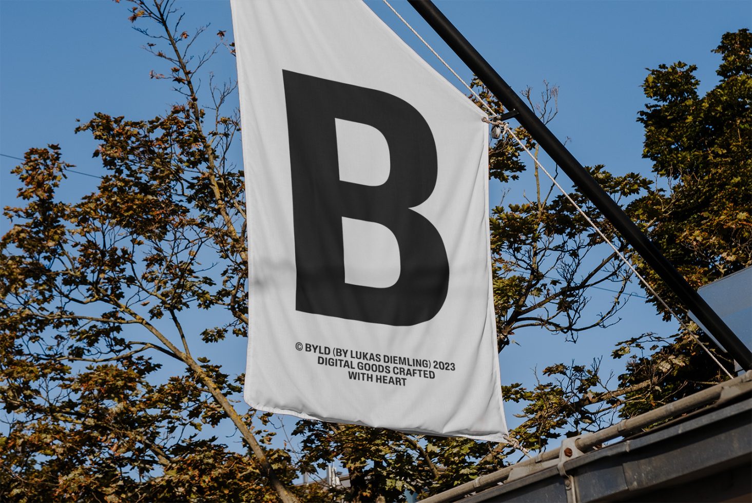 White flag mockup with black letter B design, hanging outdoors with clear blue sky and tree branches in background, for graphic display.