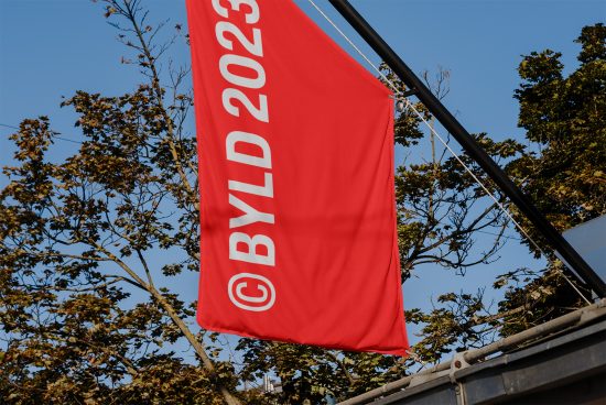 Red flag mockup with copyright symbol and text 'BYLD 2023' on pole against sky and trees, for graphic design assets.