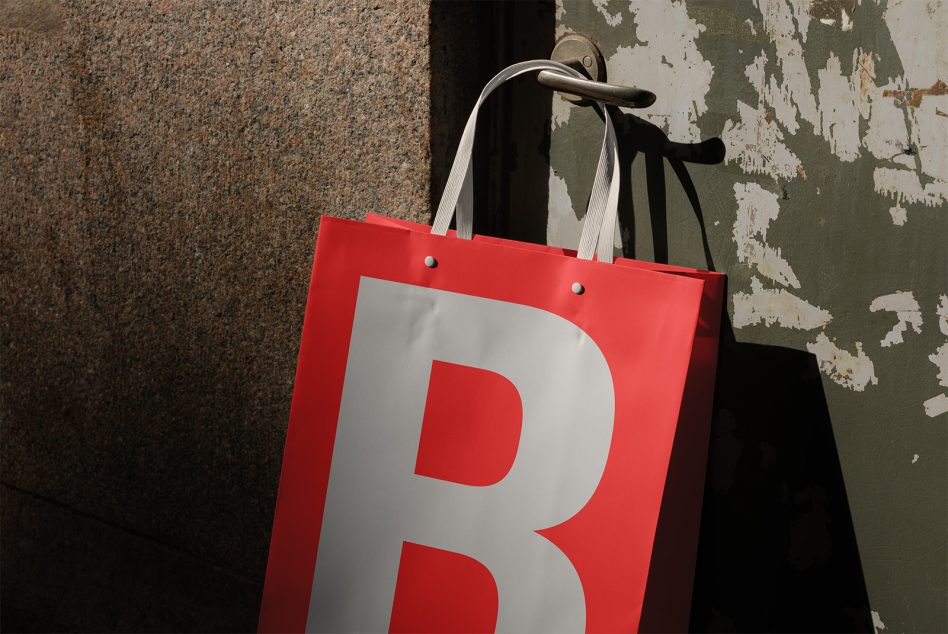 Red shopping bag mockup with bold letter B hanging on door handle, urban gritty wall backdrop, realistic shadows for product display.