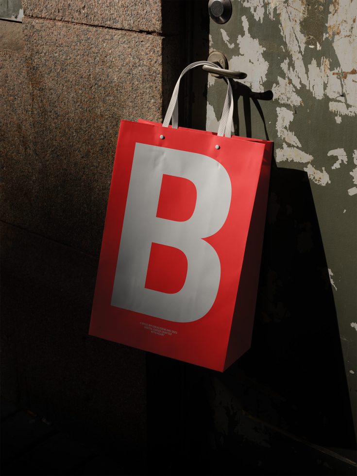 Red shopping bag with bold letter B hanging on metal handle, realistic brand presentation mockup for designers, textured background, urban style.