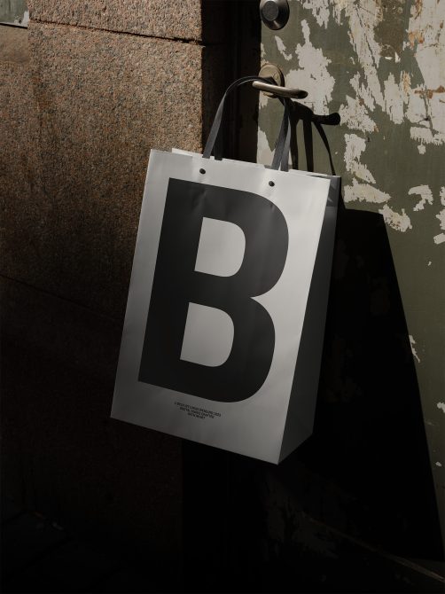 Paper shopping bag mockup with bold letter B hanging on old metal handle, urban background, ideal for branding and logo design presentation.