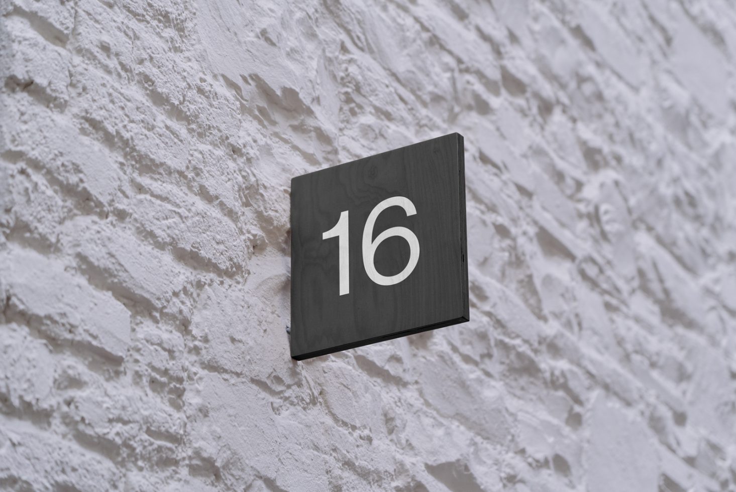 Black square number sign 16 on textured white wall, ideal for mockups and signage designers looking for minimalist presentation.