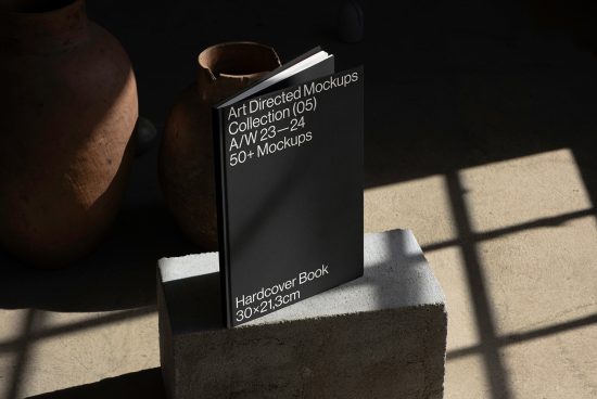Elegant hardcover book mockup on concrete pedestal with shadow play, next to rustic pottery, ideal for presentation, digital assets for designers.