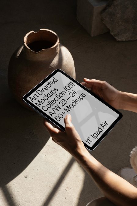 Hands holding tablet displaying Art Directed Mockups Collection ad, suggesting digital mockup template, with rustic pottery and shadows in background.