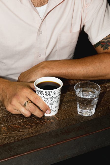 Person holding coffee cup with text, ring on finger, tattoo visible, next to glass of water on wooden table, realistic mockup for branding.