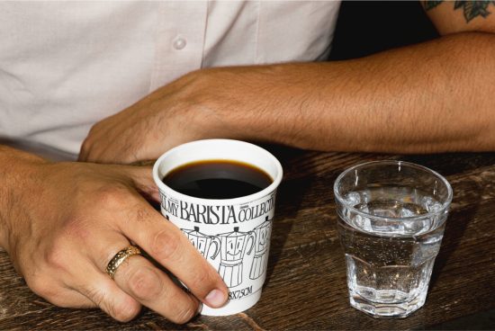 Person holding a coffee cup next to a glass of water on a wooden table, ideal for mockups with editable mug design space.