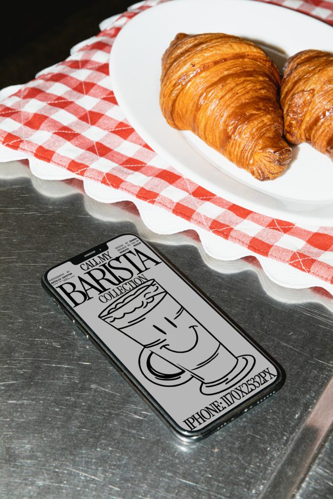 Smartphone with coffee-themed design mockup on table with croissants, perfect for designers' food-related graphics and marketing materials.