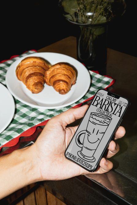 Hand holding smartphone with stylized coffee cup graphic design, croissants on a plate, cafe ambiance, digital graphics, modern mockup.