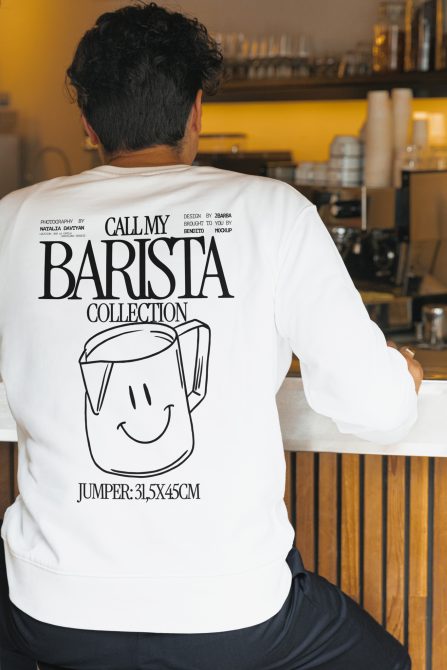 Person wearing white sweatshirt mockup with barista graphics design, posing in cafe environment, ideal for apparel presentation.