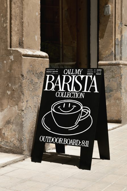 Black A-frame sidewalk signboard mockup with coffee cup design and white text, standing on a cobblestone street next to a rustic wall.