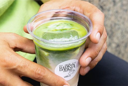 Close-up of hands holding a matcha latte cup with a label mockup on a blurred green background. Ideal for beverage branding presentation.