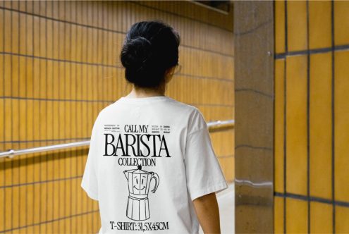 Woman standing in front of tiled wall modeling white t-shirt with barista-themed graphic design, ideal for mockup and apparel templates.