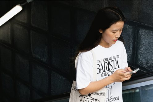 Young Asian woman in white graphic t-shirt using smartphone, urban setting, possible mockup for t-shirt design, fashion, and technology.