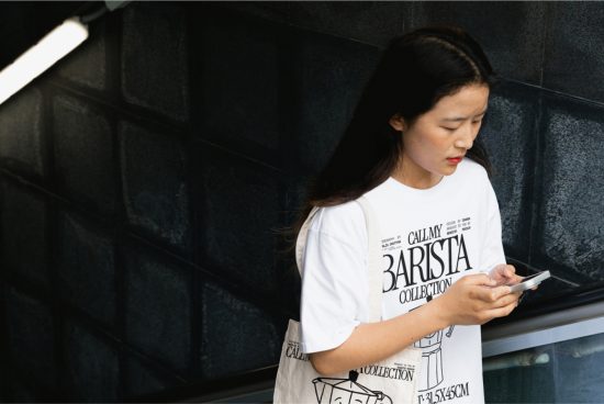 Young Asian woman in white graphic t-shirt using smartphone, urban setting, possible mockup for t-shirt design, fashion, and technology.