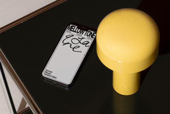 Smartphone screen mockup with stylized font showcased on dark glass table next to a modern yellow lamp, ideal for elegant branding presentation.
