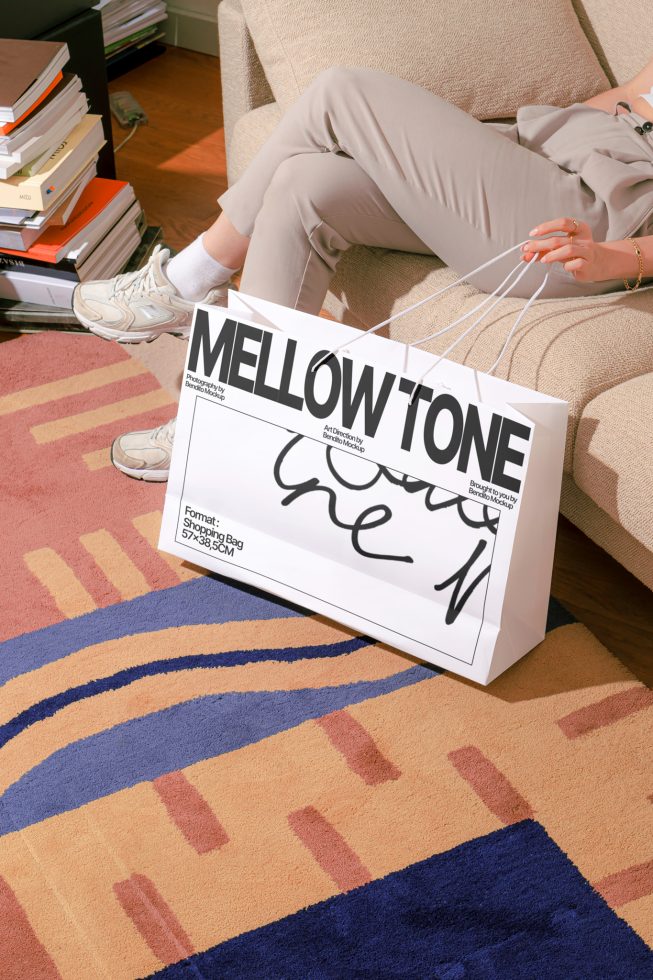 Person holding shopping bag mockup with graphic design, showcasing font style and size, in a creative indoor setting with colorful carpet.