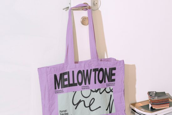 Purple tote bag mockup with bold typography hanging on a hook against a white wall with magazines nearby, ideal for designers.