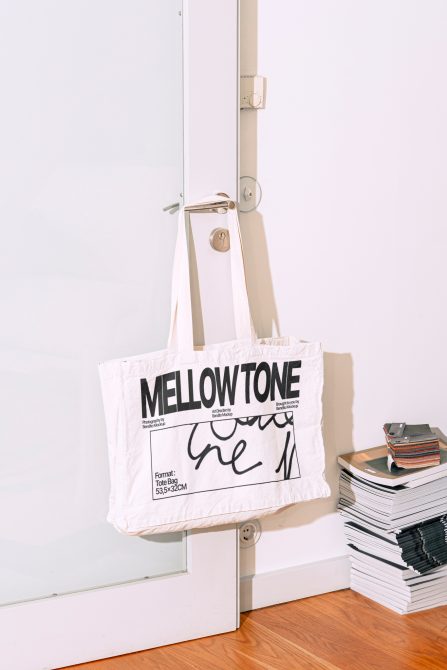 Canvas tote bag mockup hanging on a modern door handle with bold typography design, near a stack of books and vinyl records.