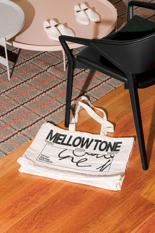 Modern tote bag mockup on wooden floor by designer chair, showcasing bold typography, ideal for presentations and portfolio displays.