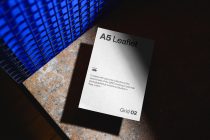 White A5 leaflet with dark text, blue grid shadow, rusty metal background