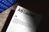 White A5 leaflet with dark text on rusty surface, blue grid light fixture visible