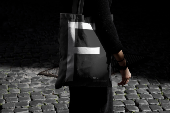 Person holding a tote bag mockup with a bold letter design on a cobblestone background for graphic design and branding presentations.