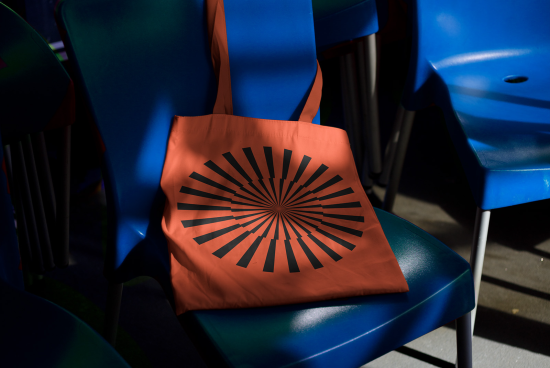 Orange tote bag with graphic black sunburst design on blue plastic chair, product mockup, stylish accessory, modern template.