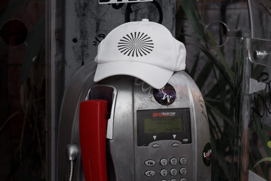 Weathered payphone wearing white cap with geometric sunburst design, red handset