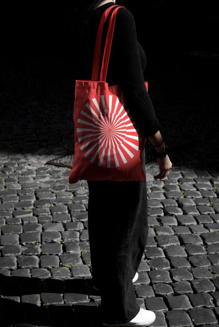 Woman standing on cobblestone street holding red tote bag with white graphic design, mockup template for designers.