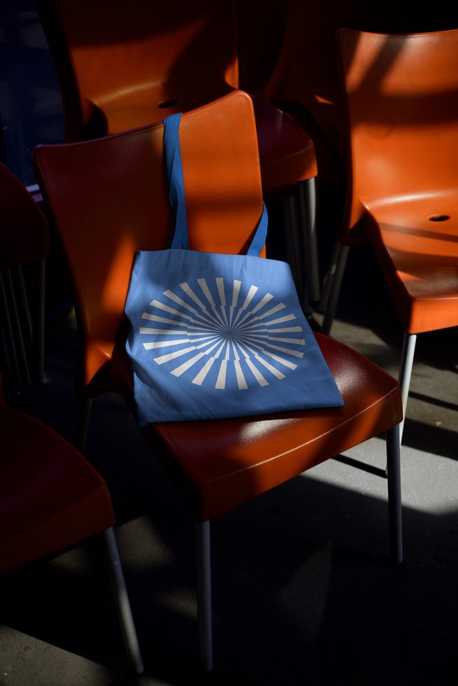 Blue tote bag with geometric design on orange chair, shadow play, product mockup for designers, creative presentation, trendy accessory.