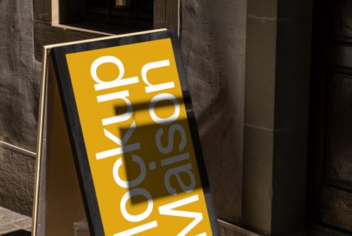 Urban street sandwich board mockup with bold yellow design, showcasing modern font style against a textured building backdrop.