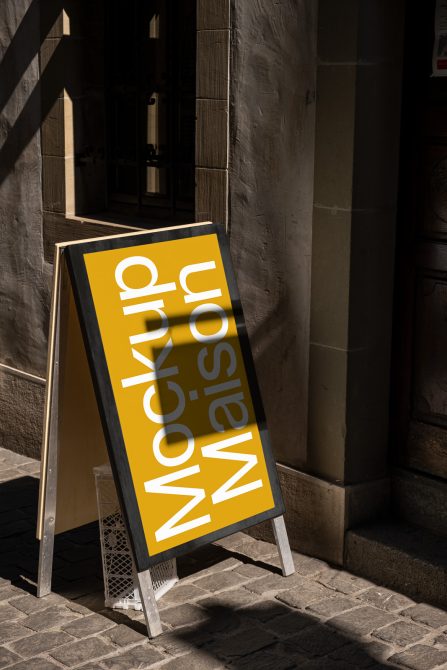 Yellow A-frame sidewalk sign mockup with customizable design, standing in sunlight with urban shadow backdrop, perfect for display ads and branding.