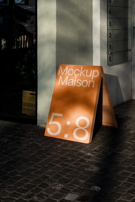 Orange outdoor mockup sign on cobblestone street next to modern building, showcasing design mockups for urban settings, shadow play.