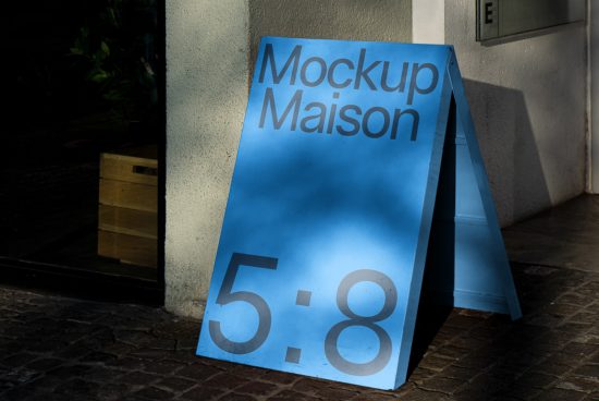 Blue A-frame signboard mockup with Mockup Maison text on a cobblestone street, under sunlight and shadows, for storefront display design.