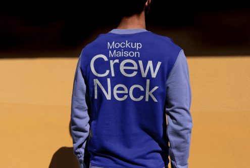 Back view of a person wearing a blue crew neck sweatshirt with mockup text for apparel design showcase against an orange wall.