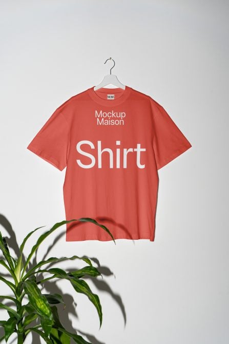 Coral red tee on hanger with white text, plant leaf in corner