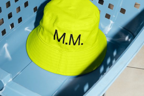 Neon yellow bucket hat with black initials M.M. on light blue crate, sunlight casting shadows, fashion mockup design asset.