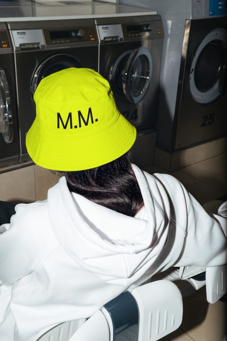 Person in white hoodie and vibrant yellow bucket hat with initials M.M. in a laundromat setting, ideal for mockup designs and fashion templates.