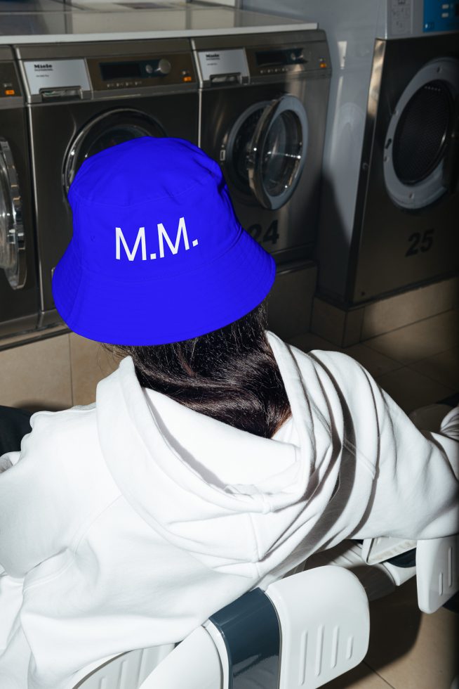 Person in white hoodie with blue bucket hat labeled 'M.M.' in a laundromat, for apparel mockup graphics design.