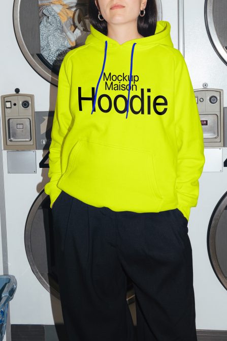 Woman in a vibrant neon yellow hoodie with mockup text, standing in a laundromat, perfect for apparel design mockups.