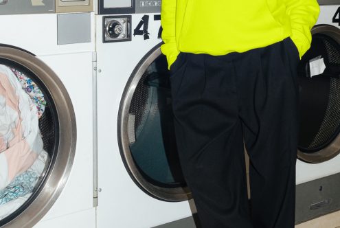 Person in bright yellow top and dark pants with hand in pocket, standing by laundry machines in laundromat. Perfect for lifestyle mockups.