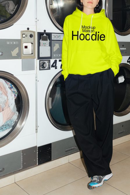 Bright yellow hoodie mockup on a model in a laundromat setting, ideal for fashion designers and apparel mockups.