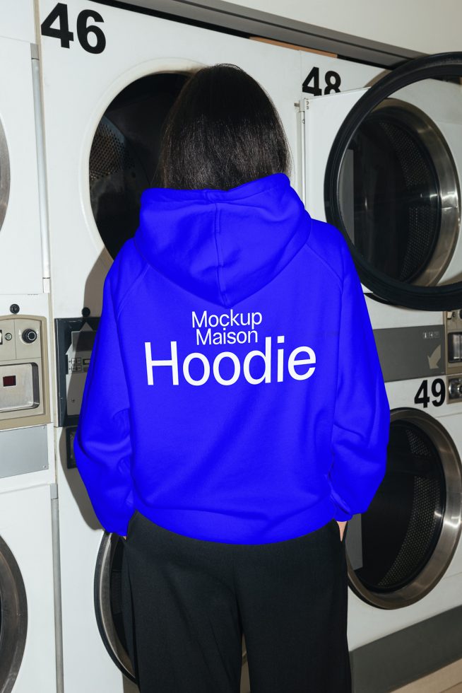 Person in vibrant blue hoodie mockup with text design, standing in a laundromat setting, perfect for showcasing apparel designs.