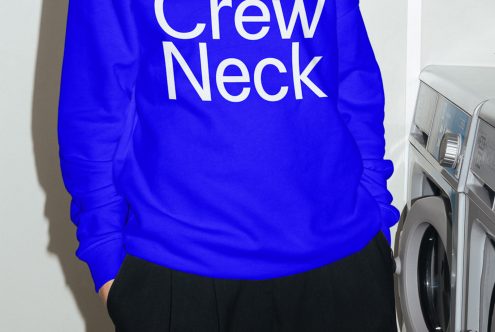 Person wearing a blue crew neck sweater mockup, white text on fabric, in front of washing machines, ideal for apparel design presentation.